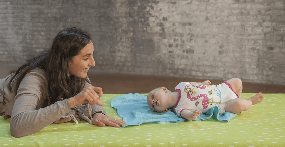 女性と赤ちゃんが、マットが惹かれた床に寝転がっている。女性は赤ちゃんに笑いかけ、赤ちゃんは女性を見つめている。